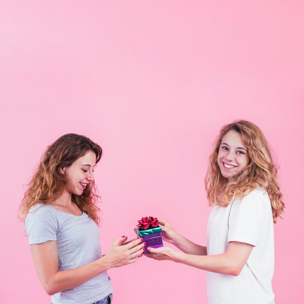 Smiling young woman giving gift to her female friend against pink backdrop