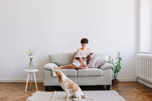 Smiling young woman in glasses looking with smile at beagle dog sitting on carpet beside sofa