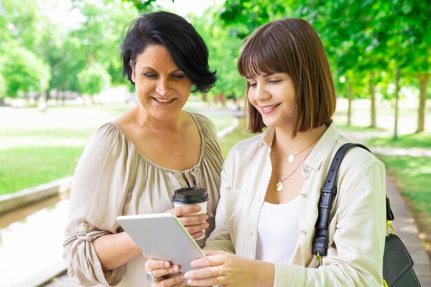 Улыбается молодая женщина и ее мать, с помощью планшета в парке