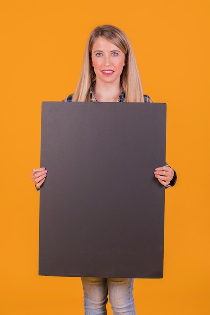 Smiling young woman holding blank black placard in hand looking at camera against orange backdrop