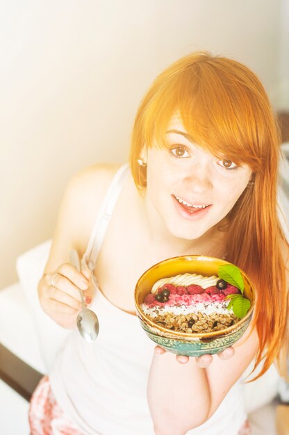 Smiling young woman holding bowl with banana; granola and fresh berries