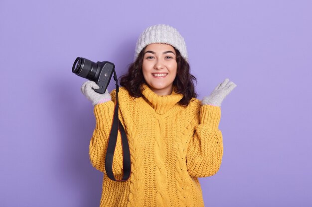 Free photo smiling young woman holding camera in one hand and spreading other palm aside