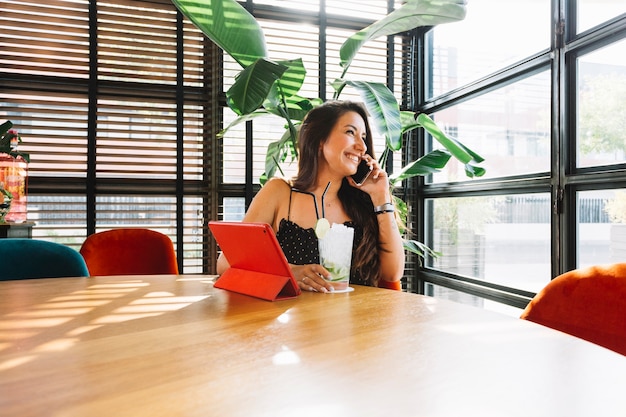 Smiling young woman holding glass of cocktail talking on smart phone in restaurant