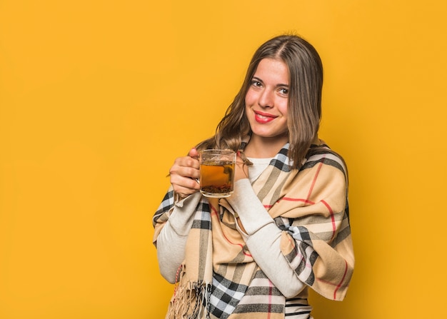 Smiling young woman holding herbal tea cup in the hands against yellow background