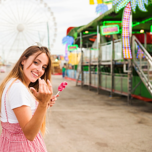 Smiling young woman holding lollipop inviting someone to come at amusement park