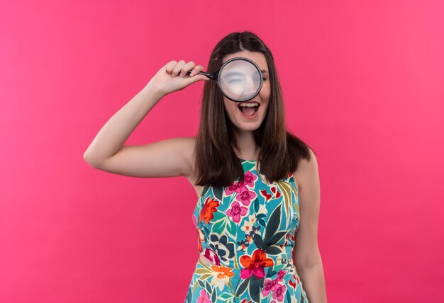 Smiling young woman holding magnifier glass and looking through it on isolated pink wall