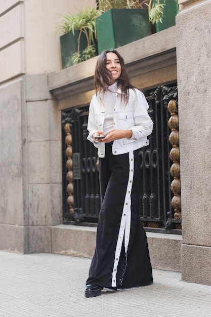 Smiling young woman holding mobile phone in hand standing on sidewalk