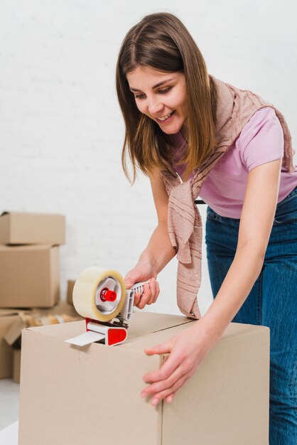 Smiling young woman holding packing machine and sealing cardboard boxes with duct tape