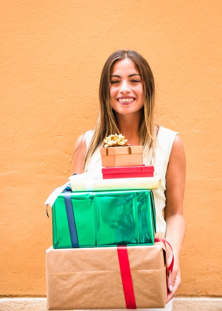 Smiling young woman holding stack of gifts against orange wall