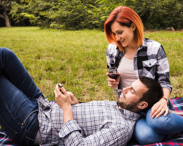 Foto gratuita vetro di vino sorridente della tenuta della giovane donna che esamina uomo che utilizza telefono cellulare nel parco