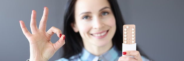Smiling young woman holds birth control pills in her hand and makes ok gesture preventing