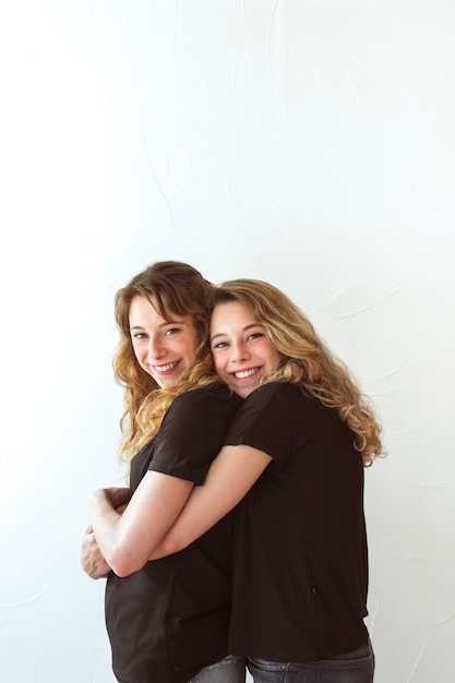 Бесплатное фото Улыбается молодая женщина, обнимает ее сестру сзади на белом фоне
