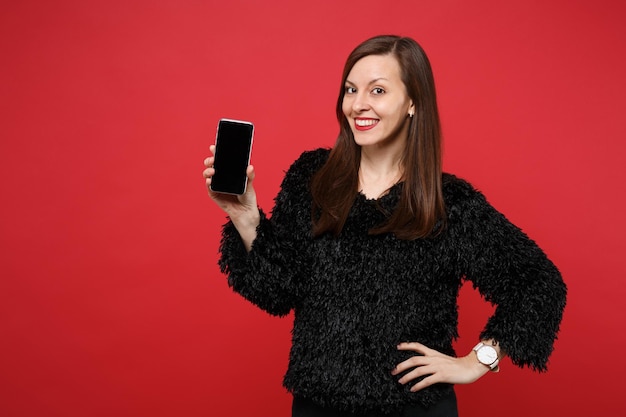 스튜디오의 밝은 빨간색 벽 배경에 격리된 빈 화면이 있는 휴대전화를 들고 검은 모피 스웨터를 입은 웃고 있는 젊은 여성. 사람들은 진심 어린 감정, 라이프 스타일 개념입니다. 복사 공간을 비웃습니다.