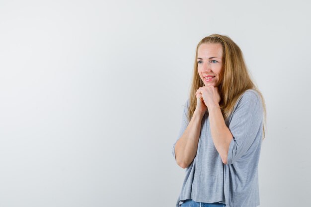 The smiling young woman is looking down by holding hands together under chin on white background