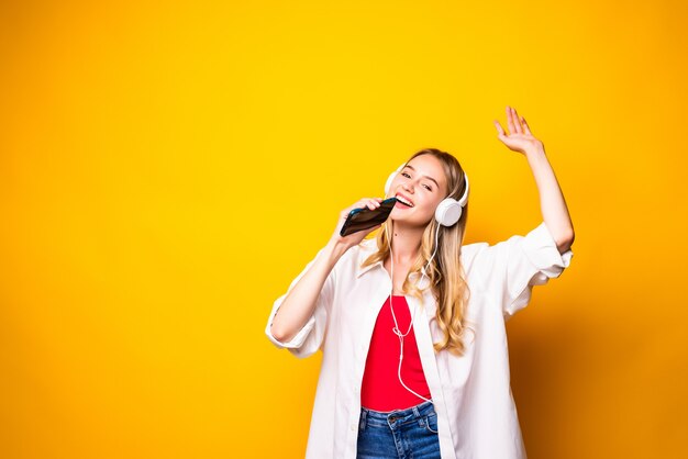 젊은 여성이 헤드폰에서 음악을 듣고 노란색 벽 위에 절연 스마트 폰을 사용하여 웃고