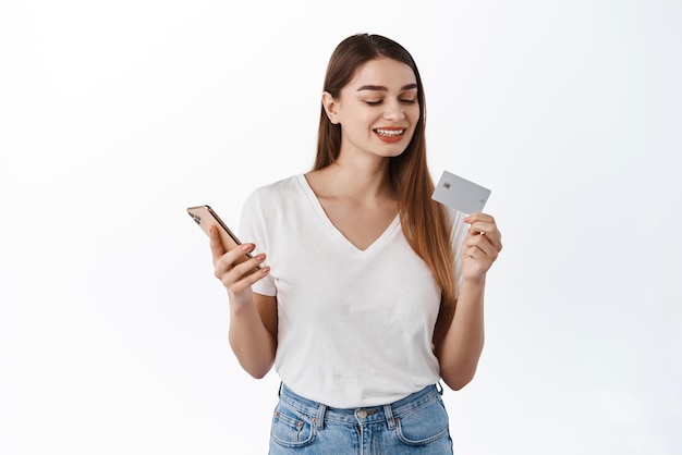 Улыбающаяся молодая женщина смотрит на номер кредитной карты во время оплаты в приложении для денежных переводов на мобильном телефоне, совершая покупки онлайн на смартфоне с помощью банковского приложения, стоящего на белом фоне
