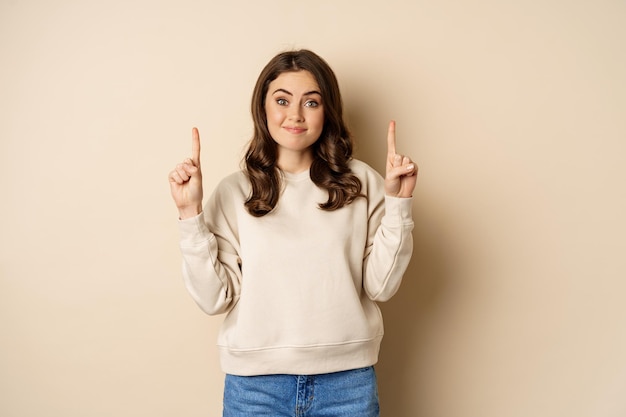 Smiling young woman looking indecisive, shrugging and smirking while pointing fingers up, standing over beige background