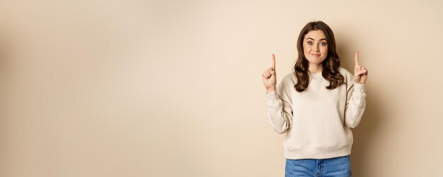 Smiling young woman looking indecisive shrugging and smirking while pointing fingers up standing ove