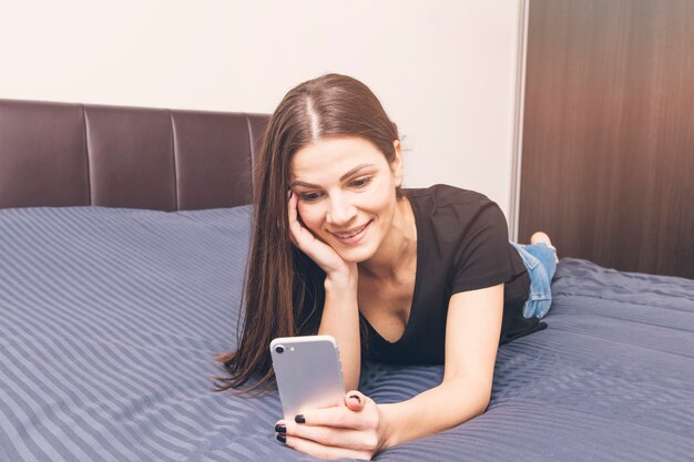 Улыбается молодая женщина, лежа на кровати, с помощью мобильного телефона