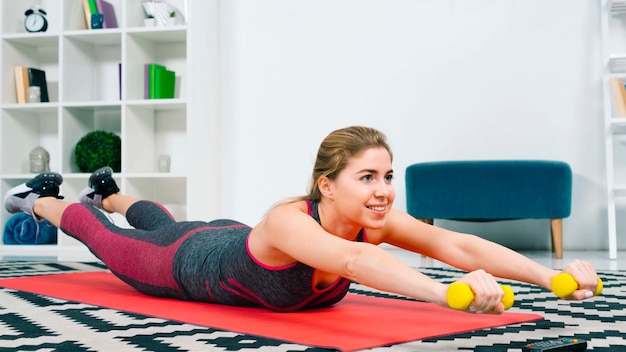 Smiling young woman lying on red exercise mat exercising with yellow dumbbells