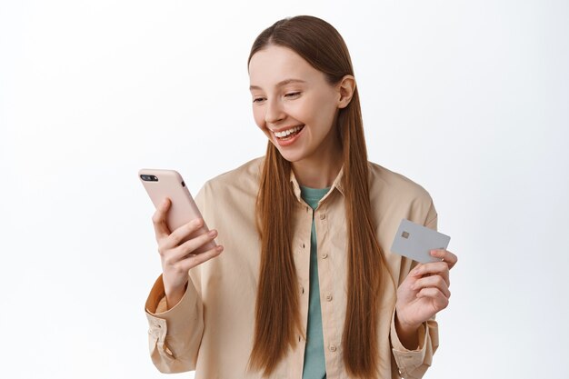 Smiling young woman make order, pay with mobile phone and credit card, looking at screen pleased, paying, standing over white wall.