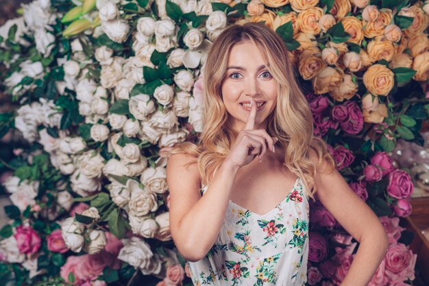 Smiling young woman making quiet gesture standing in front of roses backdrop