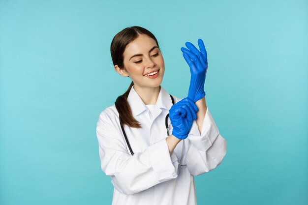 Smiling young woman medical worker wearing gloves for patient checkup standing in hospital clinic un...