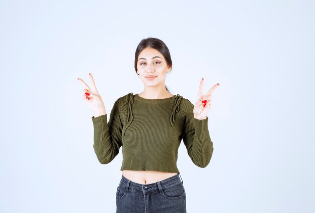 A smiling young woman model standing and showing victory sign.