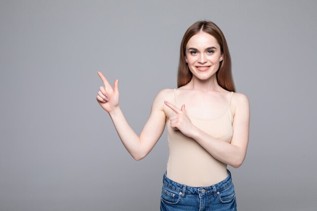 Smiling young woman pointing away and looking at the camera over gray wall