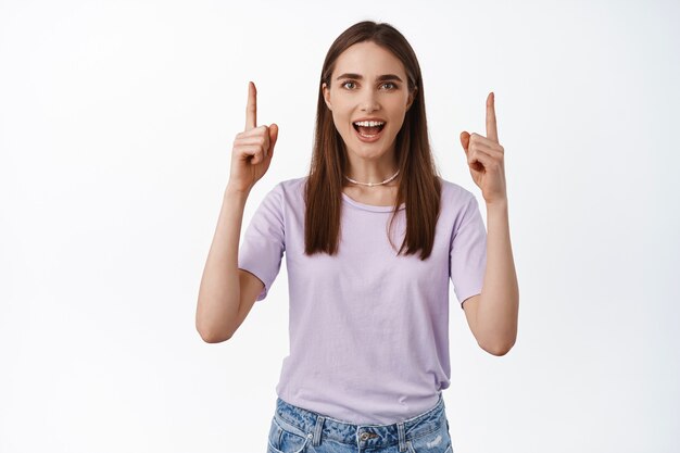 Smiling young woman pointing at place look up standing in t-shirt on white