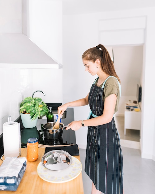 電気ストーブのソースパンで食べ物を準備している笑顔の若い女性