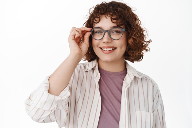 Smiling young woman put on glasses and looking at camera, standing against white background