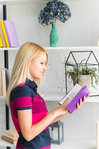 Smiling young woman reading book near shelf at home