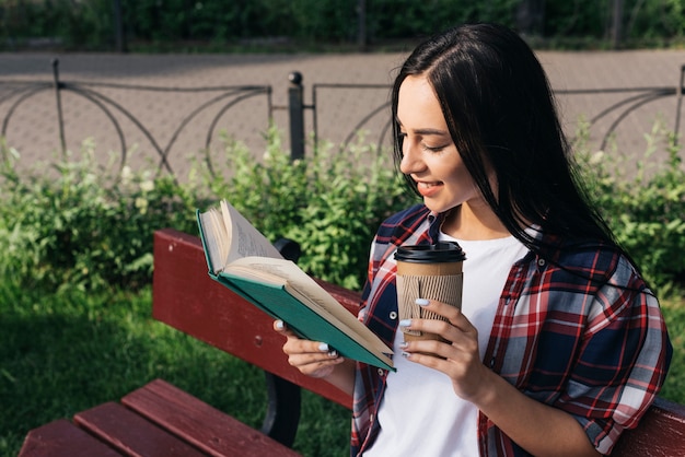 Foto gratuita libro di lettura sorridente della giovane donna con la tenuta della tazza di caffè eliminabile mentre sedendosi sul banco al parco