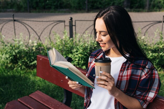 Усмехаясь книга чтения молодой женщины с держать устранимую кофейную чашку пока сидящ на стенде на парке