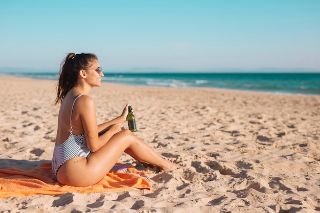 Улыбается молодая женщина, отдыхая на пляже с пивом