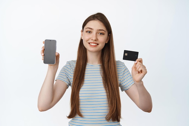 Smiling young woman showing phone screen and credit card, mobile banking and shopping concept, white background
