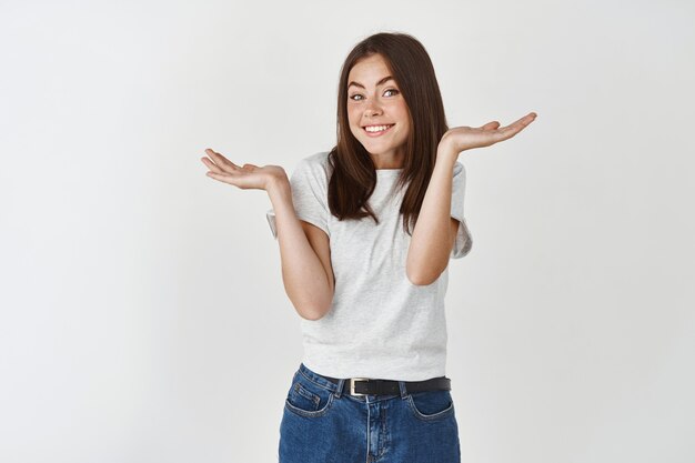 Smiling young woman shrugging shoulders and asking what, acting clueless and innocent, standing over white wall