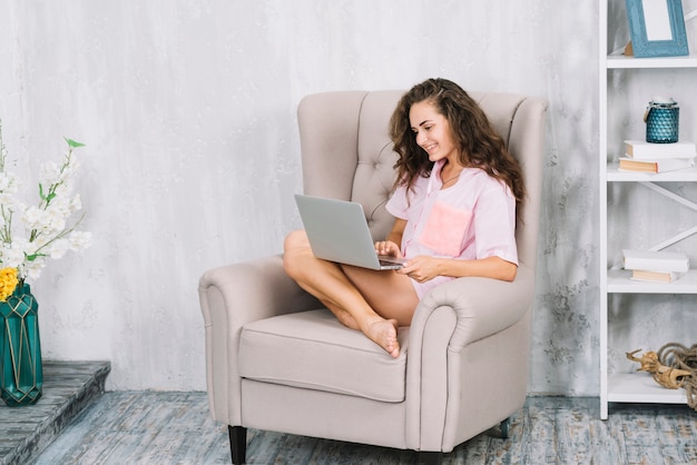 Smiling young woman sitting on armchair using laptop