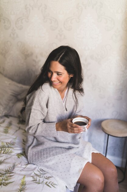Улыбается молодая женщина, сидя на кровати, держа чашку кофе