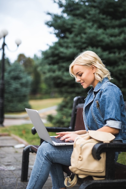 Улыбающаяся молодая женщина сидит на скамейке и использует телефон и ноутбук в городское осеннее утро