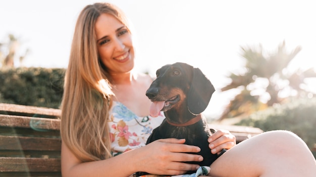 Smiling young woman sitting on bench with her dog sticking out tongue