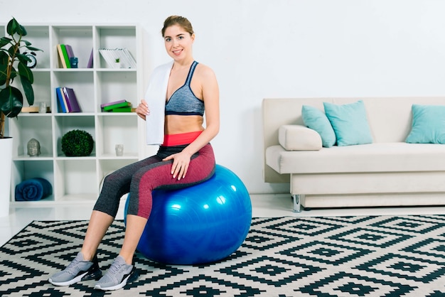 Smiling young woman sitting on blue pilates ball holding towel over shoulder
