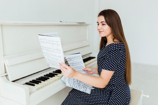 Улыбается молодая женщина, сидя перед фортепиано, глядя на музыкальный лист