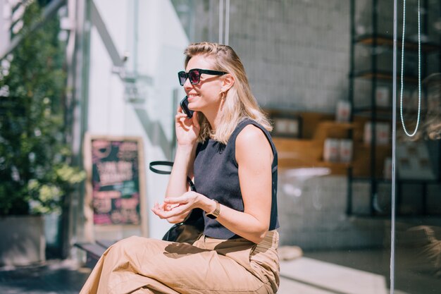 Smiling young woman sitting outside the shop talking on mobile phone