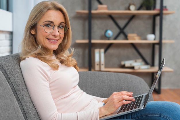 Foto gratuita giovane donna sorridente che si siede sul sofà facendo uso del computer portatile che esamina macchina fotografica