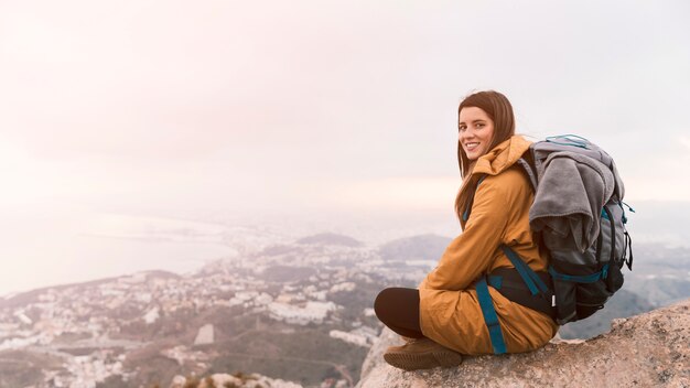 彼女のバックパックと山の上に座っている若い女性の笑みを浮かべてください。