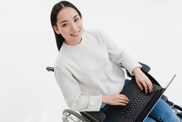 Smiling young woman sitting on wheelchair using laptop against white background