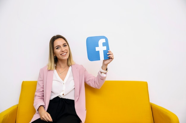 Smiling young woman sitting on yellow sofa holding facebook icon