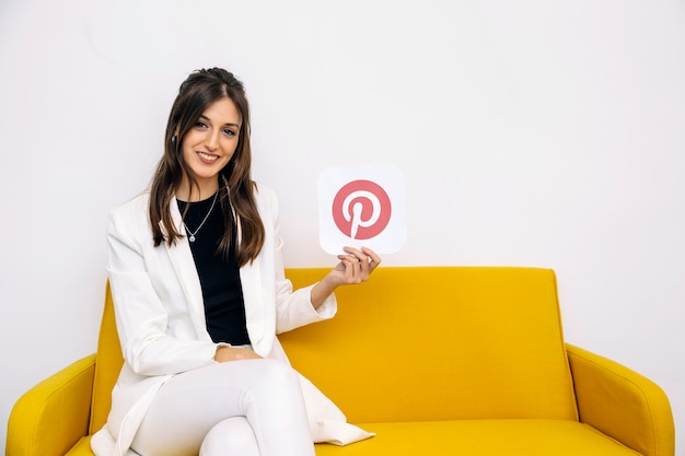 Free photo smiling young woman sitting on yellow sofa showing pinterest icon